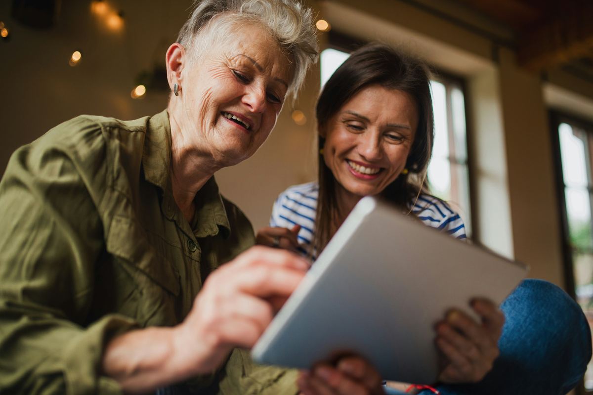 An older woman and her adult daughter smiling over a tablet at home