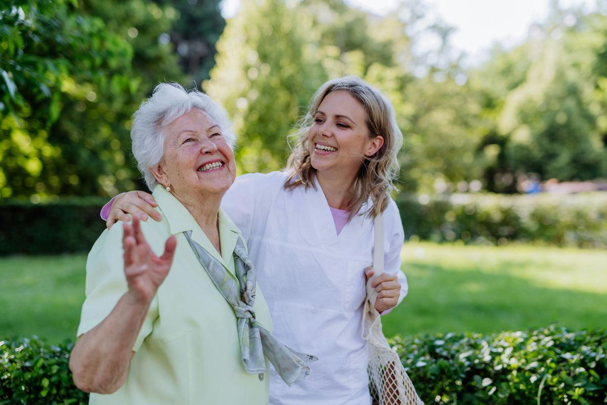 A carer with her arm around an older woman, both laughing on a walk in the park