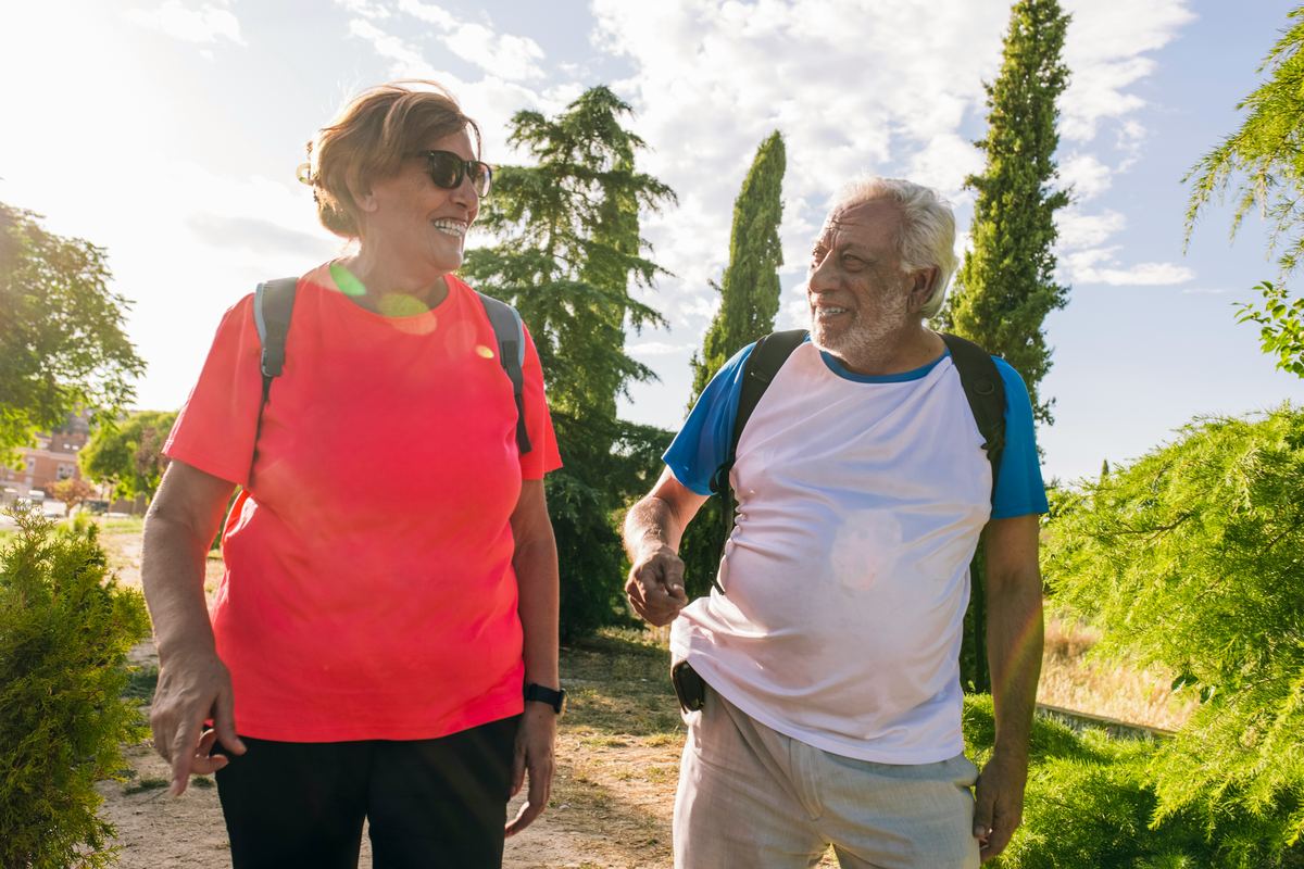 An older couple laughing as they walk together on a sunny path between cypress trees
